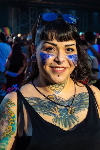 Portrait Photo of Smiling Tattooed Woman in Black Tank Top and Sunglasses with Blue Glitter on Her Cheeks Posing with Crowd of People in the Background