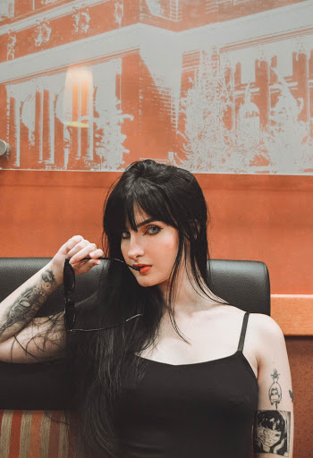 Portrait Photo of Tattooed Woman Sitting Near Orange Wall Posing With Sunglasses