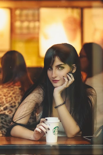 Portrait Photo of Woman Sitting Behind Window Pane With Her Head on One Hand and Holding a Starbucks Cup with the Other