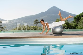 Portrait playful young woman balancing on fitness ball at sunny poolside