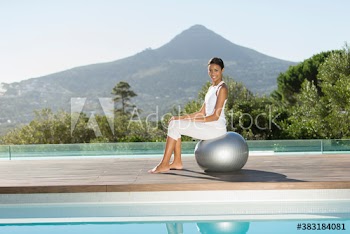 Portrait smiling young woman sitting on fitness ball at sunny poolside