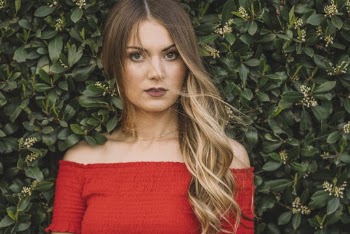 Potrait of Woman in Red Off-shoulder Top Standing Infront of Green-leafed Hedge