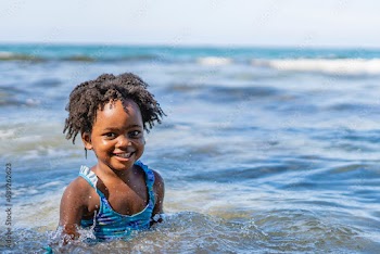 Retrato horizontal de una hermosa niña afroamericana con cabello afro muy sonriente disfrutando de un día de verano jugando con las olas el mar. 