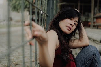 Selective Focus Close-up Photo of Woman in Red Sleeveless Top Sitting Against Metal Fence Posing
