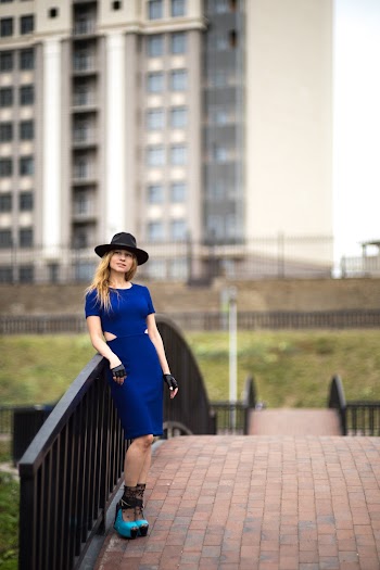 Selective focus photo of a woman in blue dress and a hat leaning on metal railings
