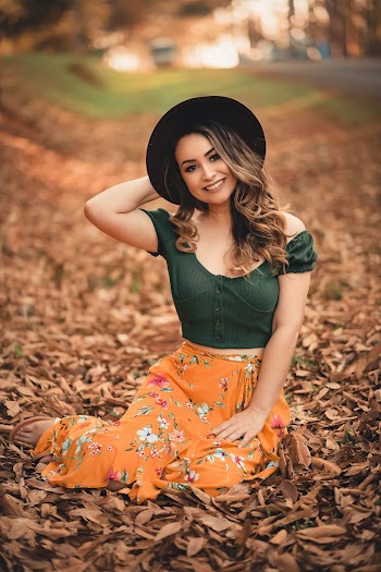 Selective Focus Photo of Smiling Woman in Black Fedora Hat, Green Top, and Yellow Floral Skirt Sitting on Dry