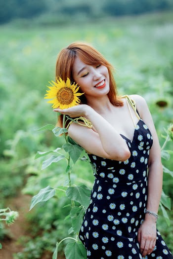 Selective Focus Photo of Smiling Woman in Black Floral Dress Posing Next to Sunflower With Her Eyes Closed
