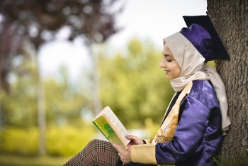 Selective Focus Photo of Smiling Woman in Hijab and Academic Regalia Sitting by Tree Reading Book