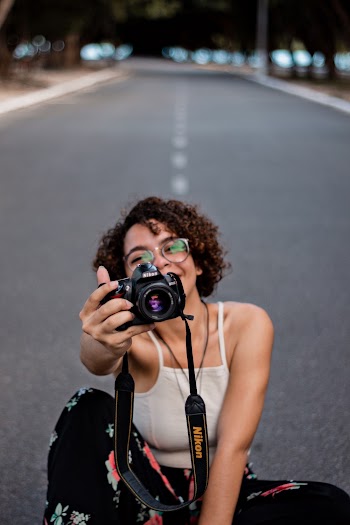 Selective Focus Photo of Smiling Woman Sitting in the Middle of the Road Holding out a Nikon Dslr Camera