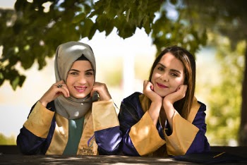Selective Focus Photo of Smiling Women in Academic Regalia Leaning on Wooden Railing With Their Hands on Their Chins