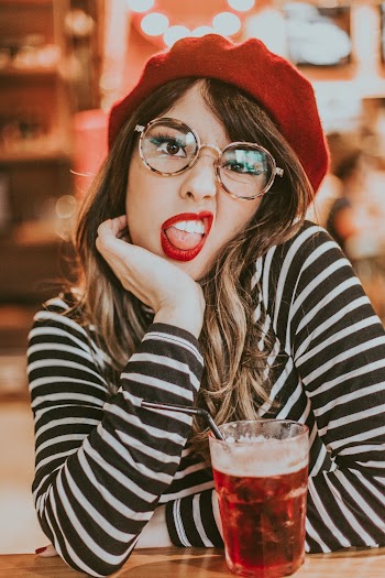 Selective Focus Photo of Woman in Black and White Striped Long-sleeved Shirt, Eyeglasses, and Red Hat Posing Next to Beverage With Her Tongue Out