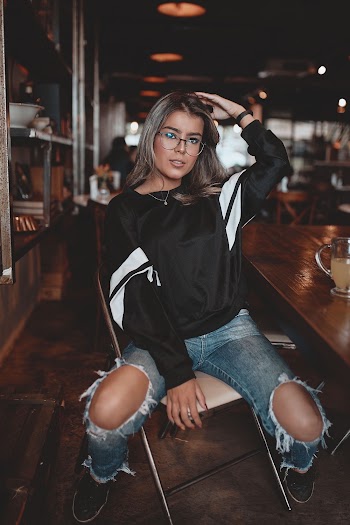 Selective Focus Photo of Woman in Black and White Sweatshirt and Blue Distressed Jeans Sitting on Steel Folding Chair Next to Wooden Table in Cafe