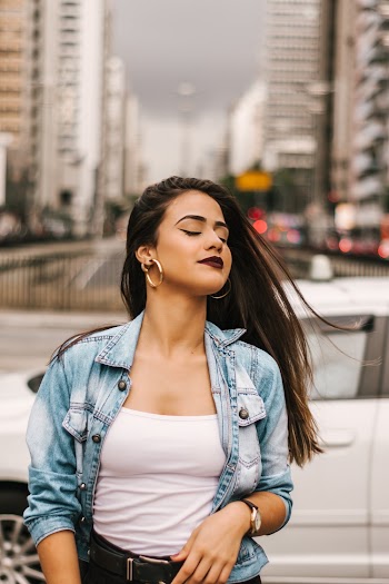 Selective Focus Photo of Woman in Blue Denim Jacket, White Top, and Black Bottoms Posing In Front of White Car With Her Eyes Closed