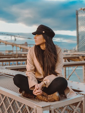 Selective Focus Photo of Woman in Brown Long-sleeved Shirt and Black Cap Sitting on Metal Surface While Looking Away