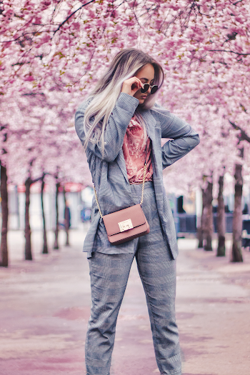 Selective Focus Photo of Woman in Gray Outfit Posing With Sunglasses Standing on Footpath Between Cherry Blossoms