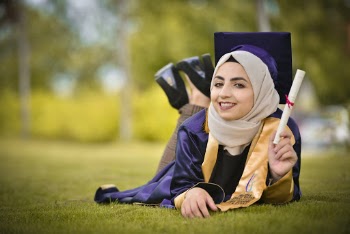 Selective Focus Photo of Woman in Hijab and Academic Regalia Lying on Grass Holding Up Diploma