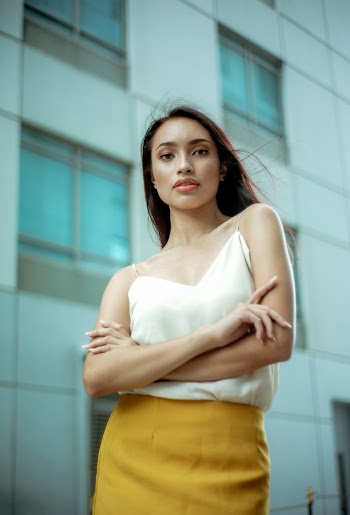 Selective Focus Photo of Woman in White Top and Yellow Skirt Posing Outside Building With Her Hands Crossed