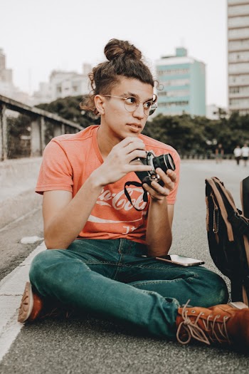 Selective Focus Photography of a Man Sitting on the Road with His Smartphone on His Lap While Holding a Black Camera