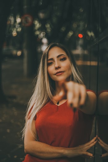 Selective Focus Portrait Photo of Woman in Red Dress Leaning on Glass Wall Posing While Pointing