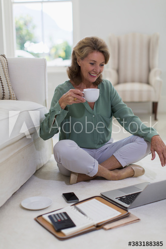 Senior woman drinking coffee at laptop on living room floor