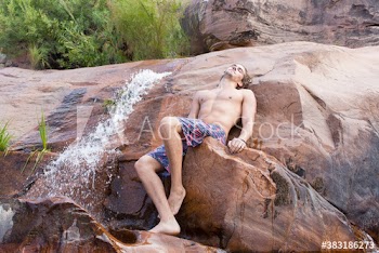Serene young man in swim trunks relaxing on rocks with waterfall