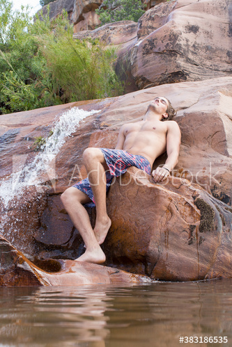 Serene young man in swim trunks relaxing on rocks with waterfall
