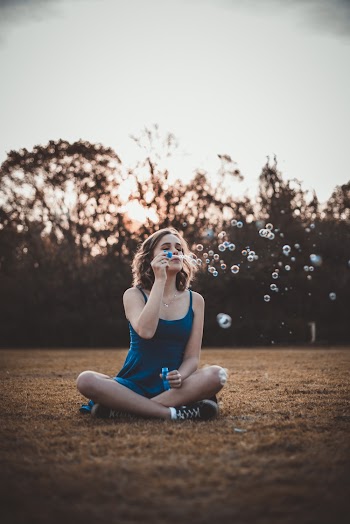 Shallow Focus Photo of Woman Sitting on Field While Blowing Bubbles
