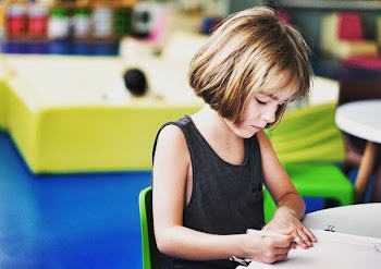 shallow focus photography of girl sitting on chair drawing on her paper on top of the table