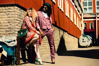Shallow Focus Photography of Woman in Orange Dress Leaning on Brown Wall Beside Man in Purple Suit Jacket
