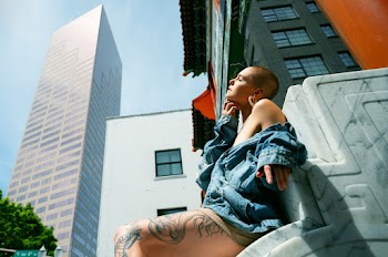 Side View Low Angle Photo of Woman in Blue Denim Jacket Sitting on Marble Surface Posing with Building in the Background