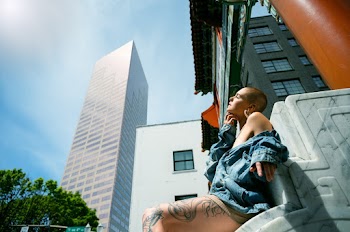 Side View Low Angle Photo of Woman in Blue Denim Jacket Sitting on Marble Surface Posing with Building in the Background