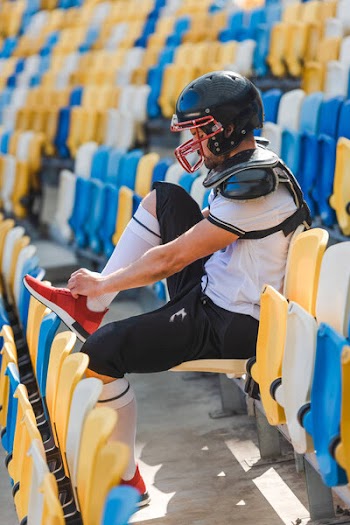 Side view of young american football player sitting on tribunes at sports stadium and lacing up shoes