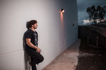 Side View Photo of Man in Black T-shirt and Jeans Holding Smartphone While Leaning on Gray Wall Posing