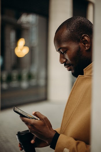 Side View Photo of Man in Brown Coat Using Smartphone While Holding Cup of Coffee