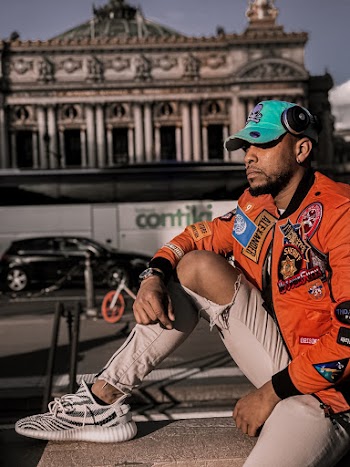 Side View Photo of Man in Orange Jacket and Brown Distressed Jeans Sitting on Stone Railing Looking  Away