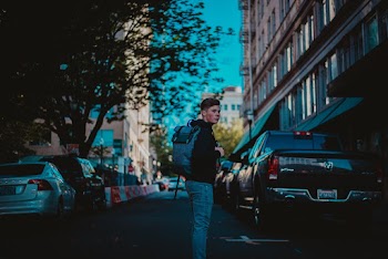 Side View Photo of Man Wearing Backpack Standing in the Middle of the Street Next to Parked Cars