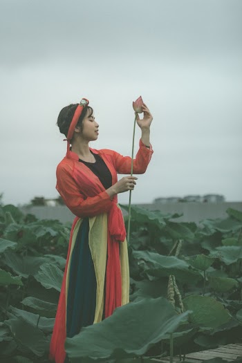 Side View Photo of Woman in Colorful Outfit Holding Pink Flower Posing with Her Eyes Closed Standing in the Middle of Flower Field
