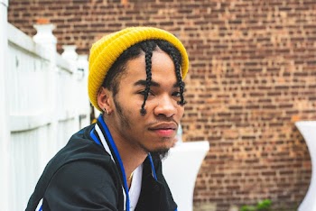 Side View Portrait Photo of Man With Braids Wearing Yellow Beanie Hat Sitting Next to Wooden White Fence