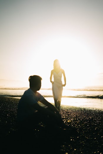 Silhouette Of Man Sitting On Seashore While Looking At Woman In White Dress Walking Towards Him