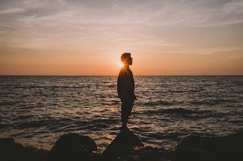 silhouette of man standing on stone beside wavy beach water during sunset