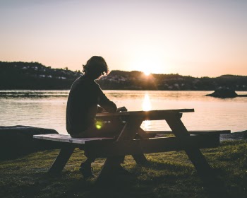 Silhouette of Person in Black Top Sitting on Picnic Bench Near Body of Water during Sunset