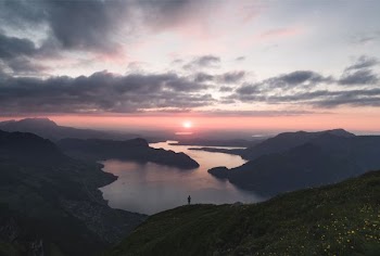 silhouette of person standing on lake surrounded by mountains under cloudy sky during golden hour