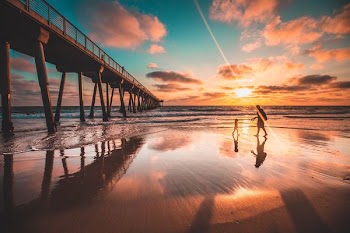 silhouette of two people walking on sand near sea beside bridge at golden hour