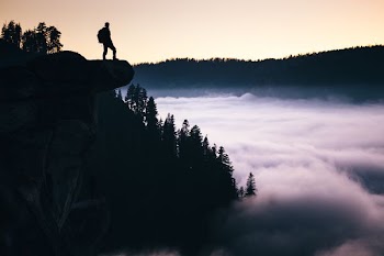 silhouette photo of man standing in mountain overlooking trees at daytime