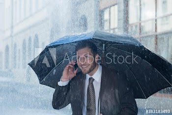 Smiling businessman with umbrella talking on smart phone in rain