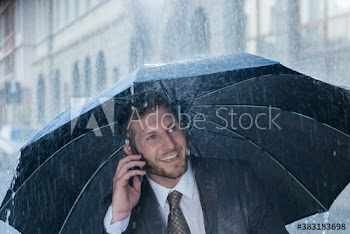 Smiling businessman with umbrella talking on smart phone in rain