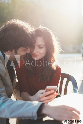 Smiling couple looking at each other while using smartphone outdoors