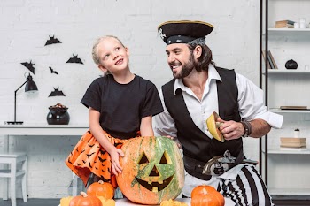 Smiling father and daughter in halloween costumes at table with pumpkin at home