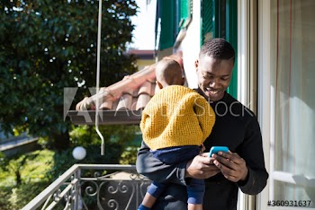 Smiling man carrying his son while using smartphone on balcony