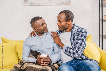 smiling senior man with cup of hot beverage sitting under plaid and looking at adult son at home